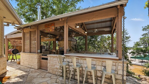 An outdoor patio with stone flooring, a wooden bar with stools, and a grill.