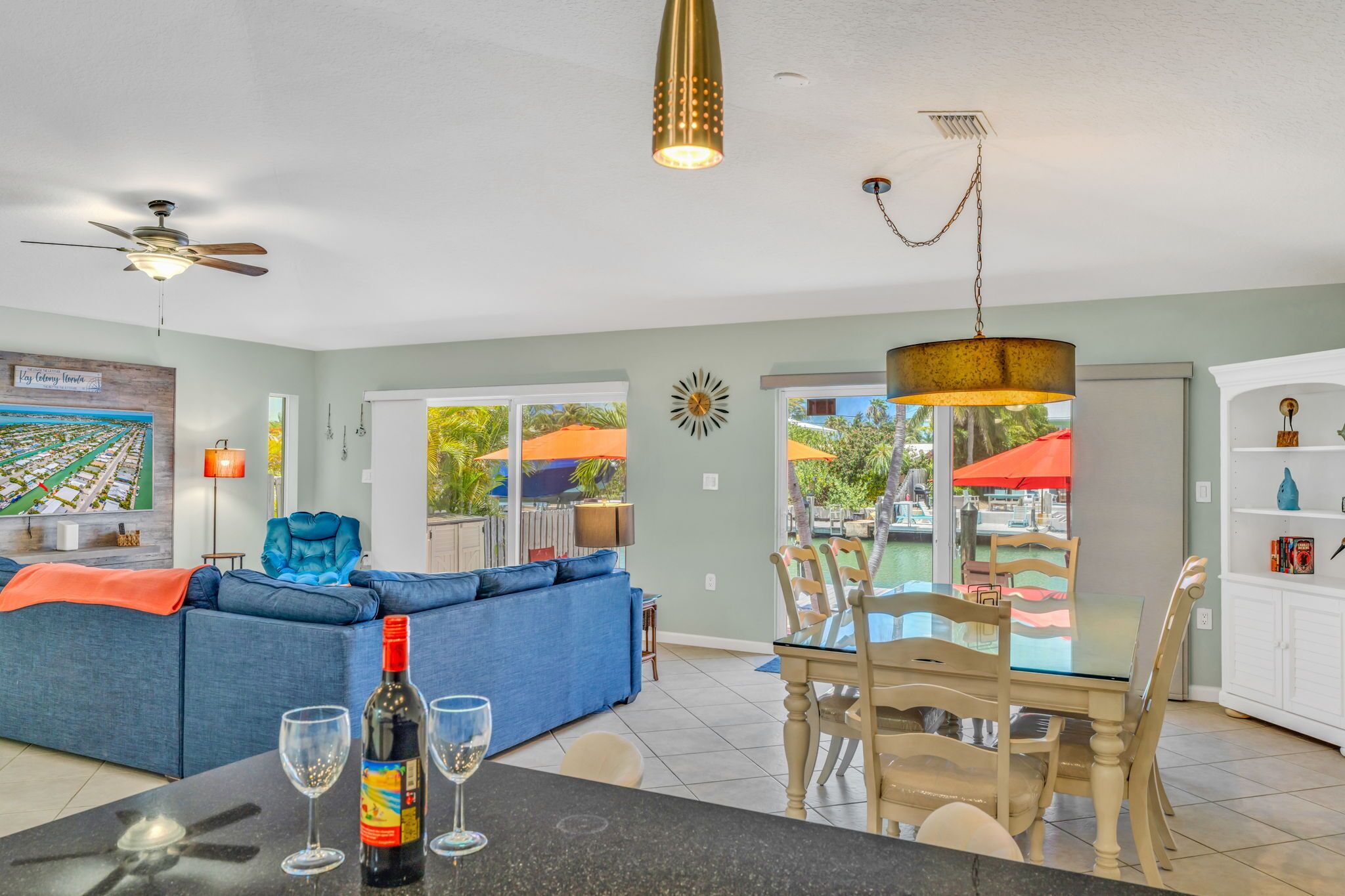 A spacious living area with a blue sofa, dining table, and chairs, a ceiling fan, and a view of the outdoors through large windows.