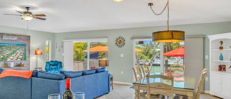 A spacious living area with a blue sofa, dining table, and chairs, a ceiling fan, and a view of the outdoors through large windows.