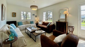 Ein Wohnzimmer mit Sofa, Sesseln, Couchtisch und einer Lampe. Zwei Fenster mit Vorhängen, eine Tür und ein an der Wand befestigter Bilderrahmen sind ebenfalls zu sehen.