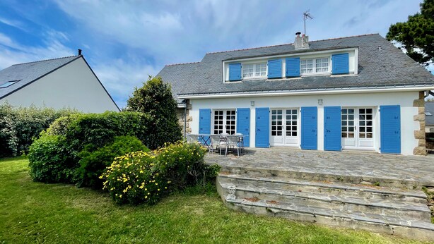 Ein zweistöckiges Haus mit blauen Fensterläden, einer Steintreppe und einer kleinen Terrasse mit Stühlen.