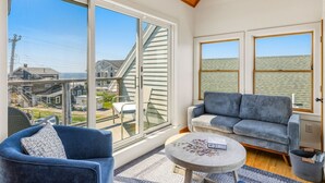 Una habitación con un gran ventanal corredero, un sofá azul, una pequeña mesa redonda con un rompecabezas y vistas a casas a través de las ventanas.