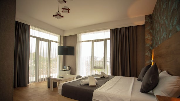 A hotel room with a large bed, a desk, a chair, and a view of the city through large windows. - Pollux (Batumi)