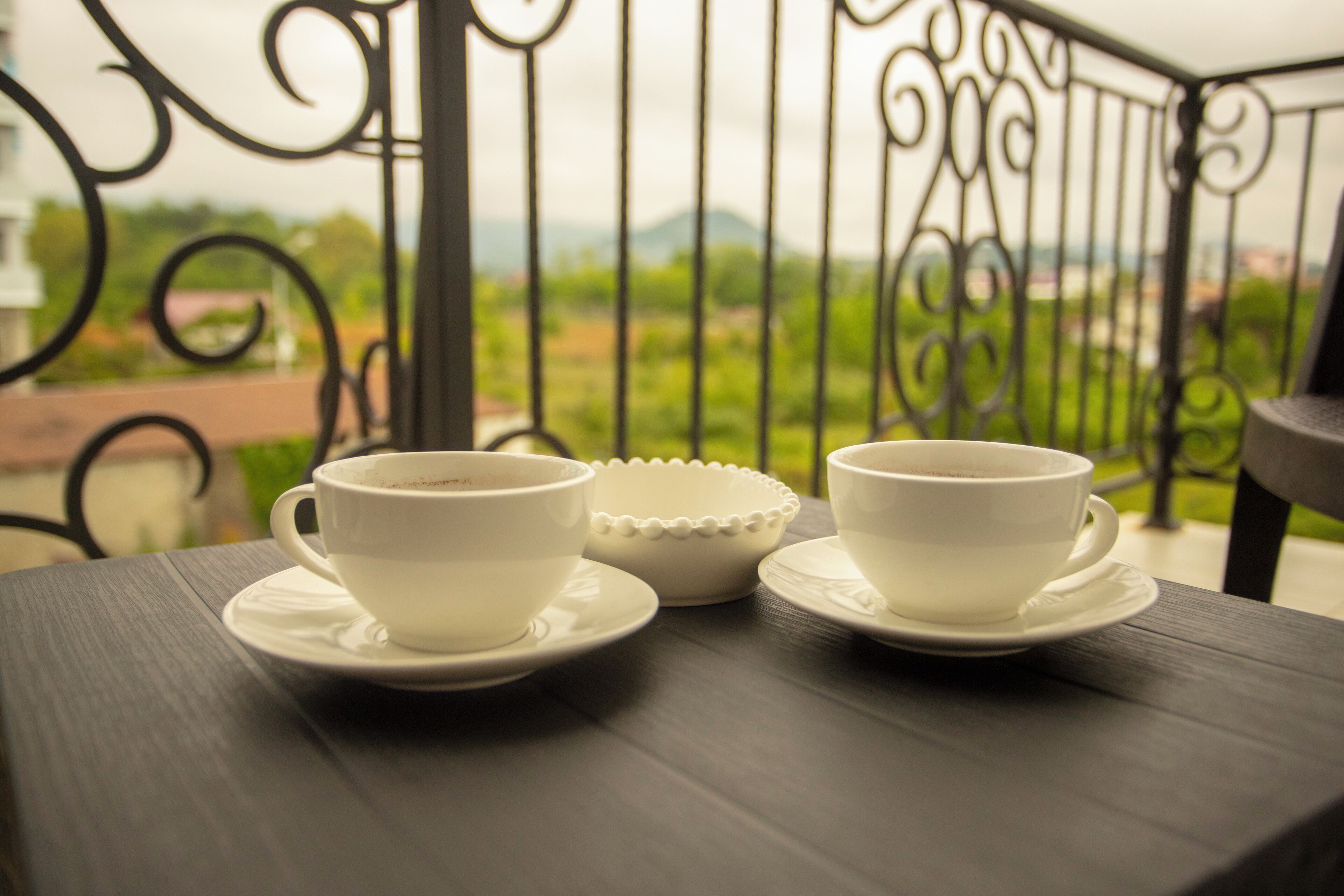 A table with two white cups and a saucer on a balcony with a metal railing and a view of greenery and buildings in the background.