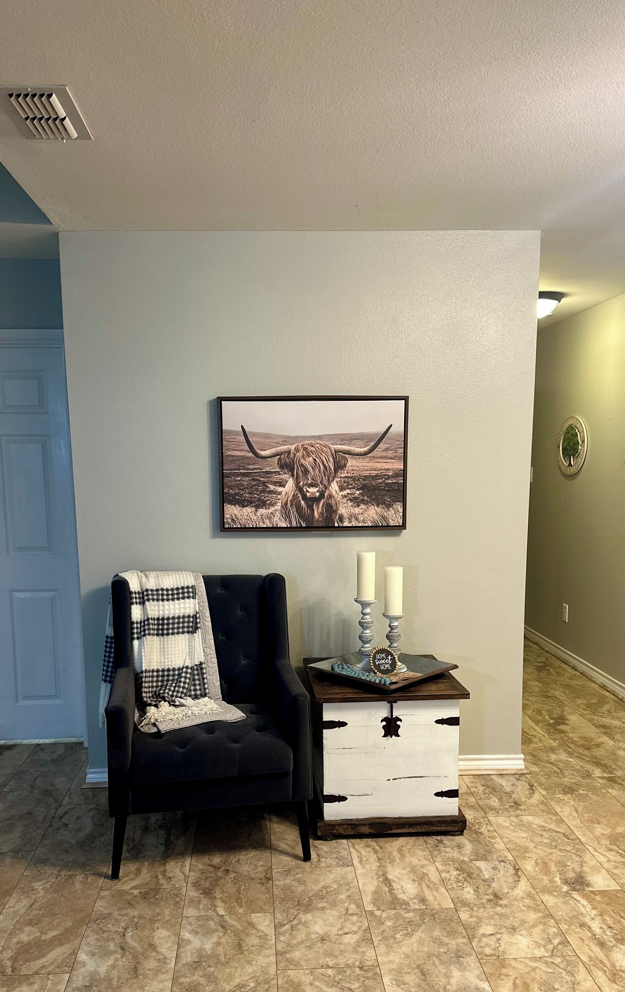 A living room with a black armchair, a white and black checkered throw blanket, a framed picture of a yak, and a side table with candles and a decorative plate.