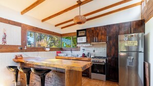 A modern kitchen with wooden beams, a central island with stools, a stainless steel refrigerator, and a window with a view of greenery. - RitoKazo (Quintero)