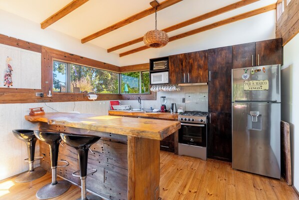 A modern kitchen with wooden beams, a central island with stools, a stainless steel refrigerator, and a window with a view of greenery.