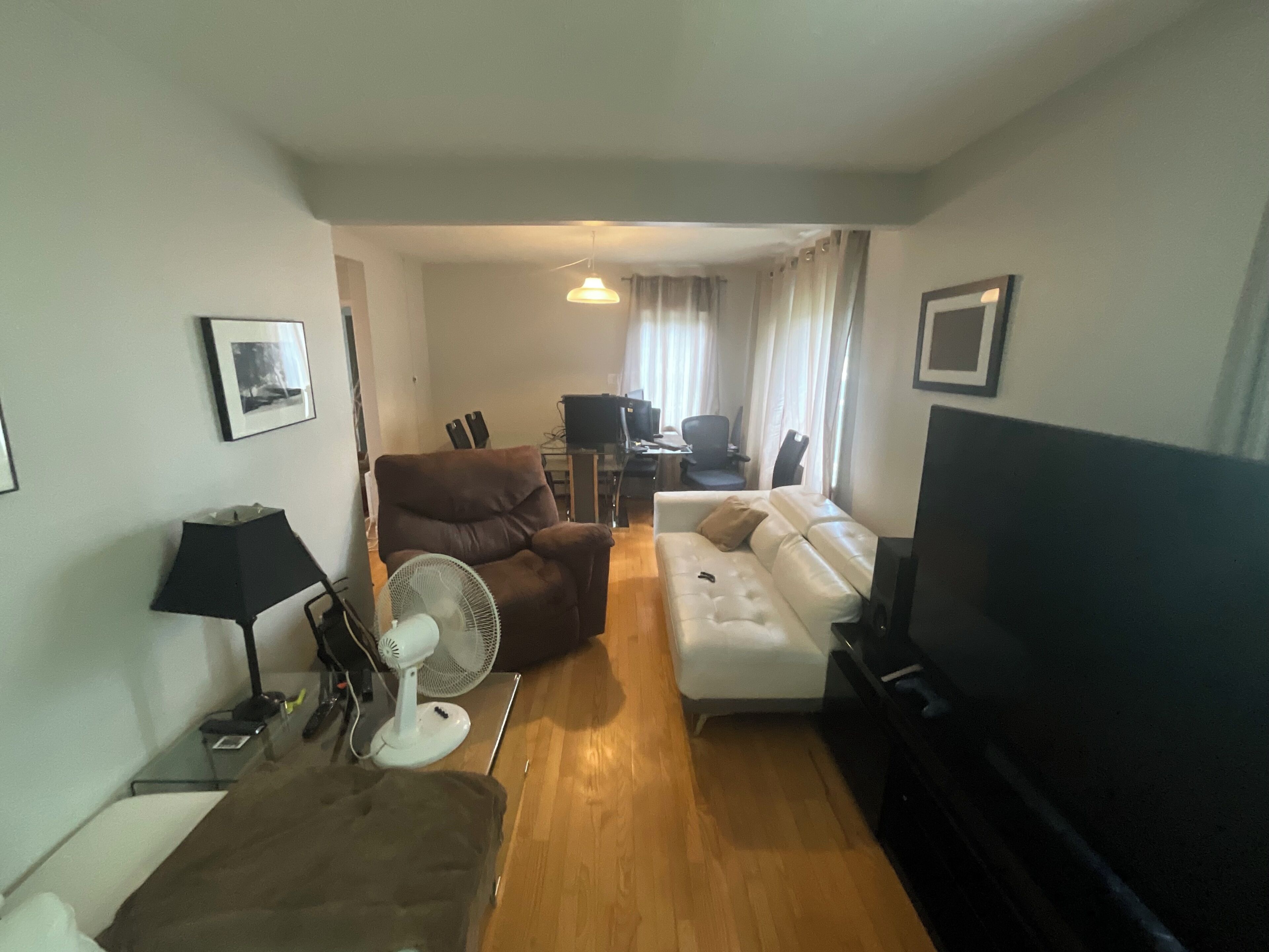 Ein Wohnzimmer mit einem braunen Ledersessel, einem weißen Sofa, einem Fernseher und einem Ventilator. Im Hintergrund steht ein Esstisch mit Stühlen.