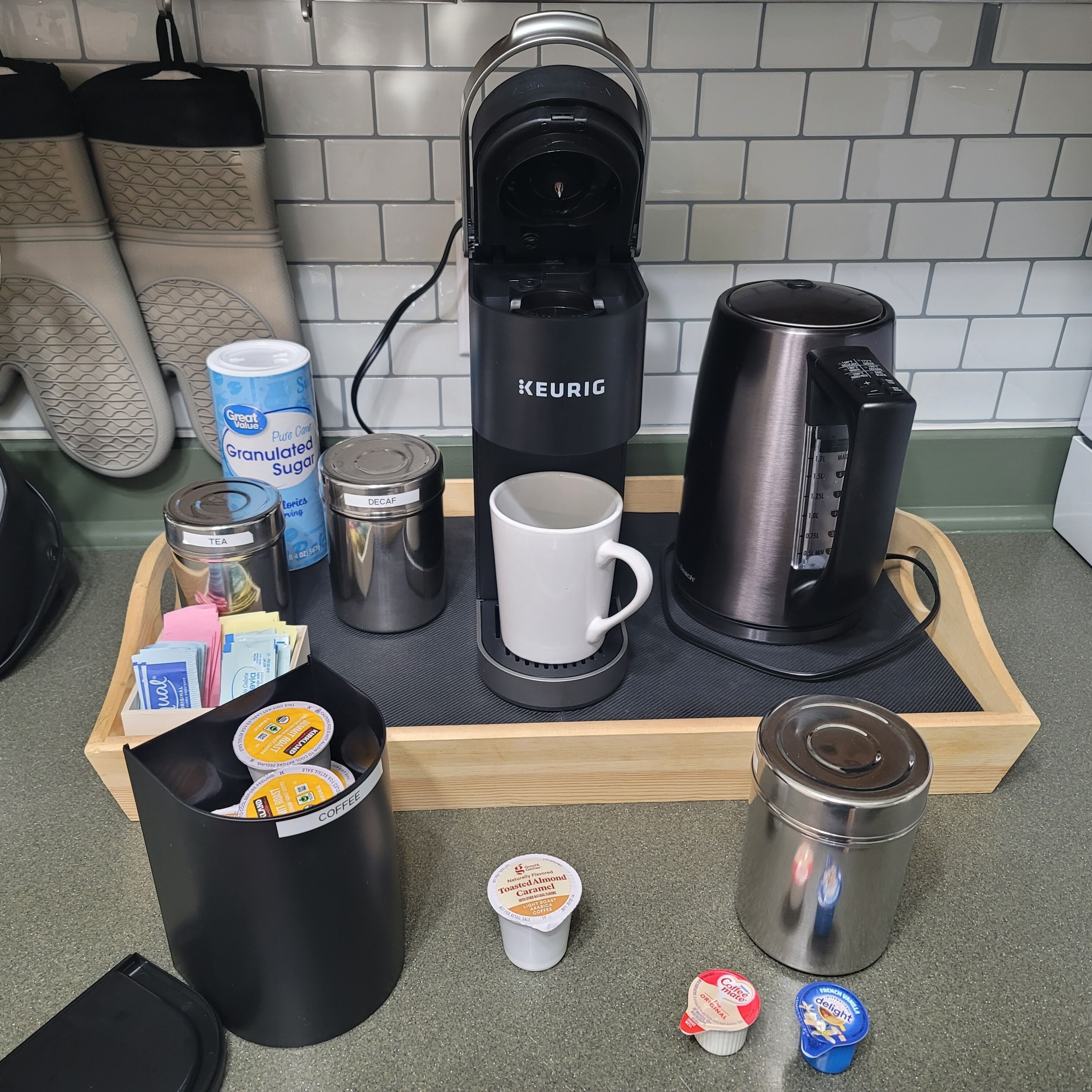 A coffee station with a Keurig coffee maker, a mug, a thermos, a coffee pod holder, a jar of sugar, and a container of creamer.