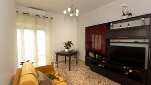 A living room with a sofa, a TV stand with decorative items, a dining table with chairs, and a window with sheer curtains. - Florence Oasis (Firenze)