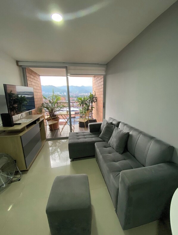 Ein modernes Wohnzimmer mit einer grauen Couch, einem Fernseher und einem Balkon mit Blick auf die Stadt und Berge.