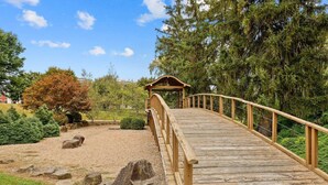 Un pont en bois menant à un belvédère situé dans un jardin aménagé, agrémenté de rochers et d’arbres.