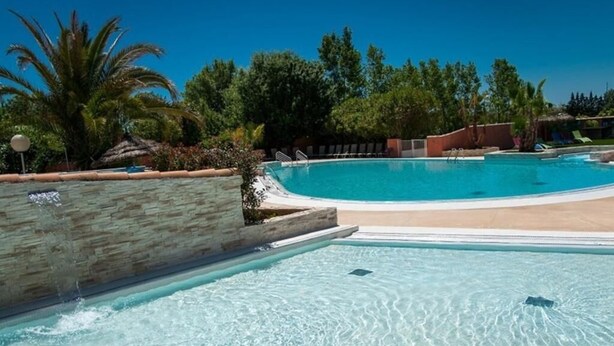 En pool med klart vatten, en stenmur på ena sidan och ett sandområde med solstolar i bakgrunden.