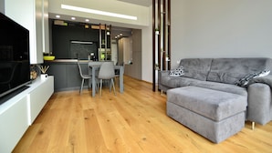A modern living room with a grey sofa, wooden flooring, and a kitchen area with a dining table and chairs. - Deluxe apartment the Chestnut (Madonna della Castagna)