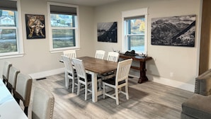 Un comedor con una mesa de madera y sillas blancas, ventanas amplias con vistas al paisaje y obras de arte en las paredes.