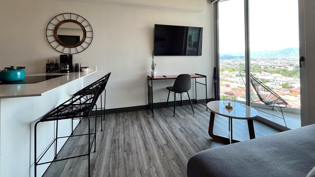 A modern hotel room with a large window, a flat-screen TV, a desk, a chair, a small round table, and a sofa. - Secrt Sabana 1BR · Workspace + WiFi · Parking · Apt 1508 (Mata Redonda)
