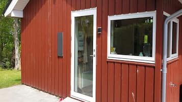 En rød bygning med dør- og vinduesramme i hvid, et synligt el-skab, samt en beton terrasse med et par sko.