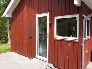 Ein rotes Gebäude mit weiß umrahmter Tür und Fenster, einem sichtbaren Stromkasten und einer Beton-Terrasse mit einem Paar Schuhen.