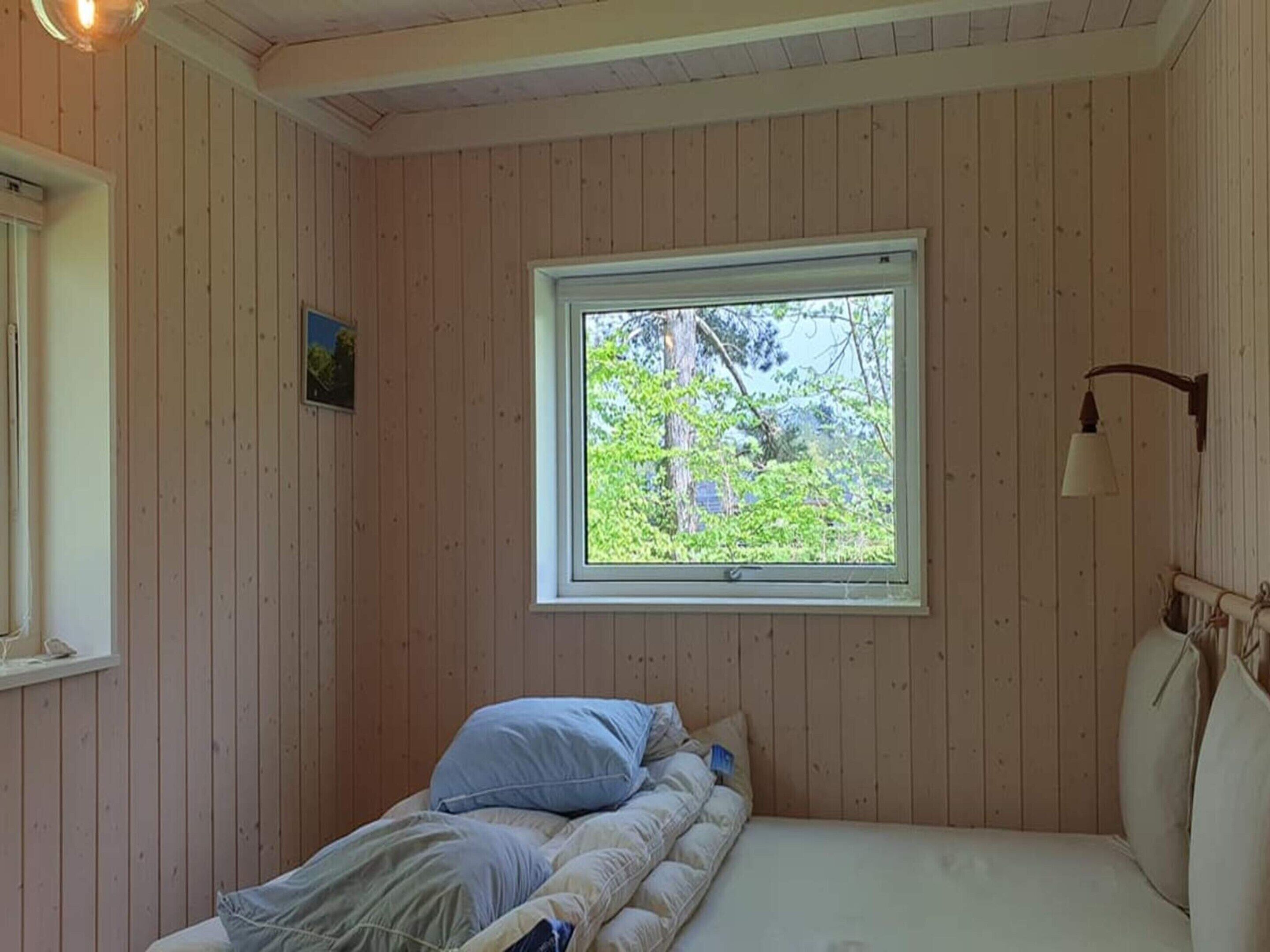 Ett sovrum med en enkel säng, ett fönster med utsikt över grönområden och en vägglampa.