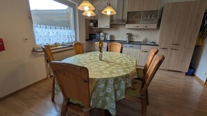 A dining area with a round table covered by a floral tablecloth, surrounded by wooden chairs. There is a window with a view of a fence and a radiator under the window.