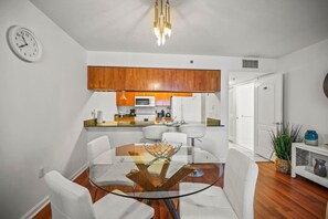 Cozinha moderna com armários de madeira, micro-ondas e cafeteira. Há uma área de jantar com mesa de vidro e cadeiras brancas.