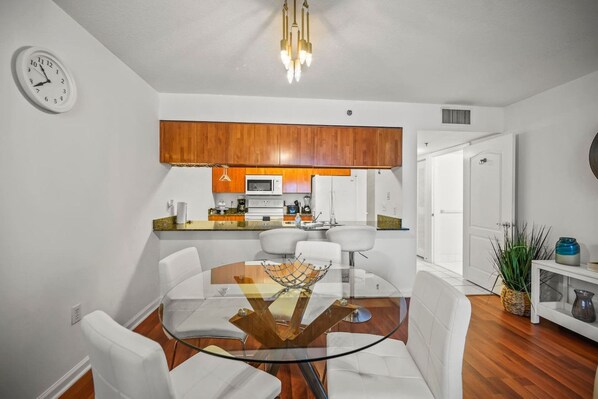 A modern kitchen with wooden cabinets, a microwave, and a coffee maker. There is a dining area with a glass table and white chairs.