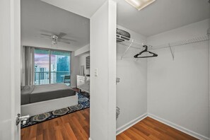 Une chambre avec un lit, un ventilateur de plafond, une fenêtre donnant sur des immeubles, une commode et un placard avec un porte-vêtements.