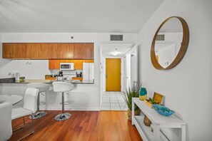 Une cuisine moderne avec des meubles en bois, un plan de travail blanc et un four à micro-ondes. On y trouve un coin repas blanc avec des tabourets de bar et une console blanche agrémentée d’objets décoratifs. Une porte en bois donne accès à une autre pièce.