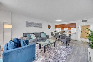 Una sala de estar moderna con un sofá gris, sillones azules y una mesa de centro de vidrio. Cuenta con un comedor con una mesa y sillas blancas, y una cocina con armarios de madera y un microondas.