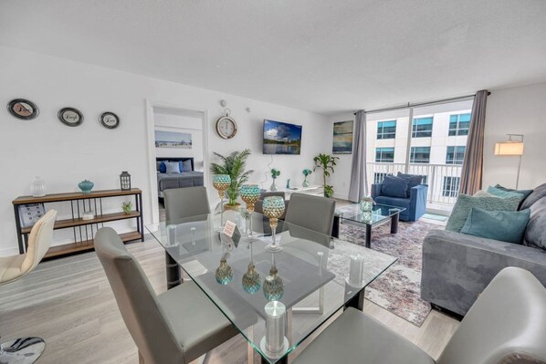 A modern living room with a glass dining table, grey sofas, and a balcony with a view.