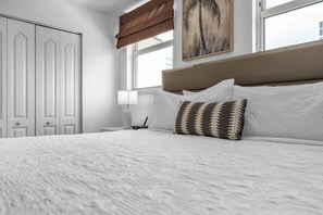 A bedroom with a large bed, white bedding, a nightstand with a lamp, a painting on the wall, and a window with blinds. - Panoramic 2BR Bay-View in Brickell With Parking (Miami)