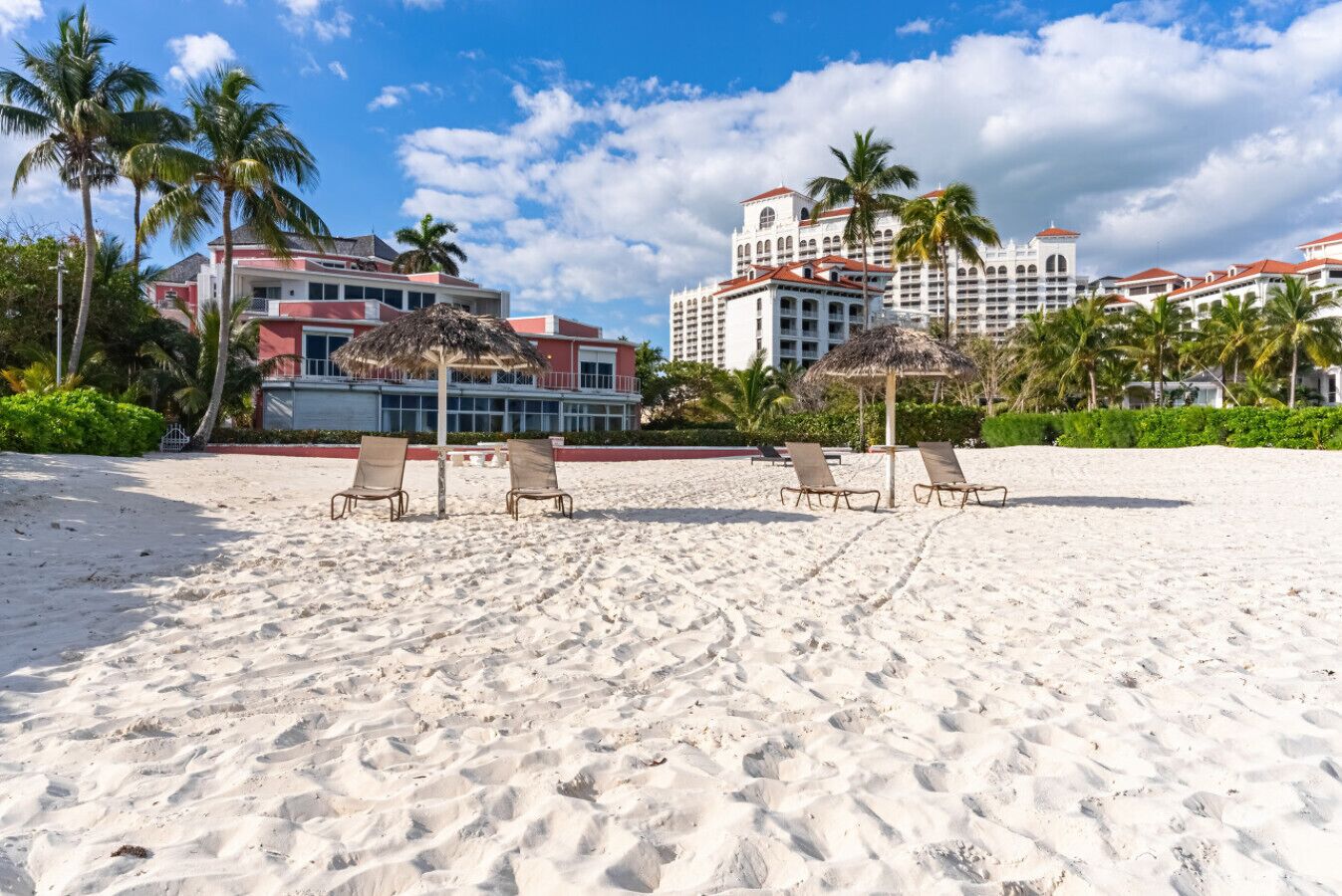 A beach with lounge chairs and a thatched umbrella, palm trees, and a large hotel building in the background.