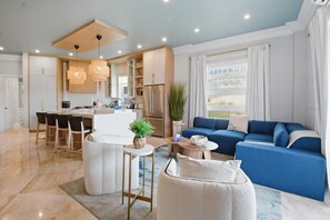 Ein modernes Wohnzimmer mit einem blauen Sofa, einem weißen Sessel und einem Essbereich, im Hintergrund eine Küche.