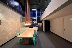 Eine moderne Küche mit einer zentralen Kochinsel, einem Essbereich mit Holztisch und türkisfarbenen Stühlen sowie einer hohen Decke mit Stadtansicht-Mural.