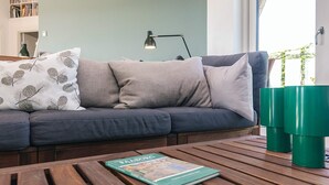 Ein modernes Wohnzimmer mit grauem Sofa, gemusterten Kissen, einem Couchtisch mit Büchern, einer grünen Lampe und Blick auf Grünflächen.