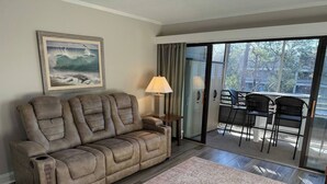 Salón con un sofá reclinable, una lámpara, un tapete estampado y vistas a un balcón con mobiliario exterior.