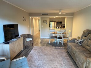 Sala de estar com televisão, sofá, poltrona, mesa de jantar com cadeiras e cozinha ao fundo.