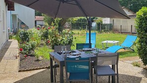 Een terras met een tafel en stoelen onder een grote parasol, omgeven door een tuin met bloemen en een hek.