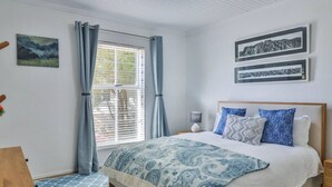 A bedroom with a bed, a nightstand, a lamp, a window with curtains, and two framed pictures on the wall.
