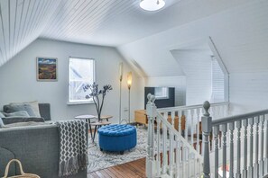 Une pièce de style « loft », avec un plafond haut, un escalier, un canapé, un pouf bleu et un tableau accroché au mur.