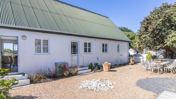 A building with a green roof, white walls, and a gravel driveway. There are outdoor chairs and a table, and a small garden area with plants and a cactus.