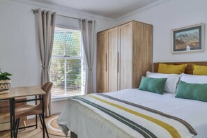 A bedroom with a bed, a desk, a chair, a window with curtains, and a painting on the wall.