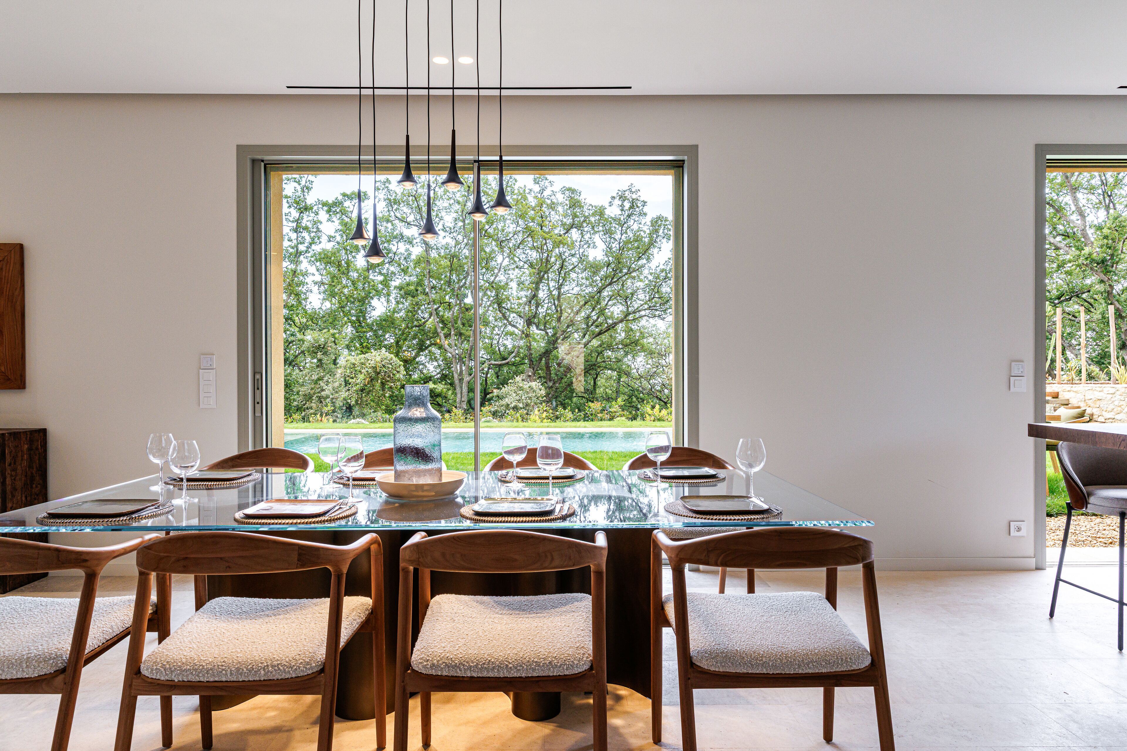 Une salle à manger moderne avec une table en verre, des chaises en bois et une grande fenêtre donnant sur un paysage arboré et une pelouse.