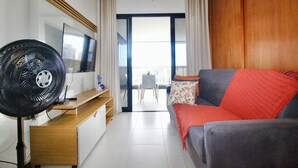 Ein modernes Hotelzimmer mit Flachbildfernseher, Standventilator, Sofa mit roter Decke und Blick auf die Stadt durch große Fenster.