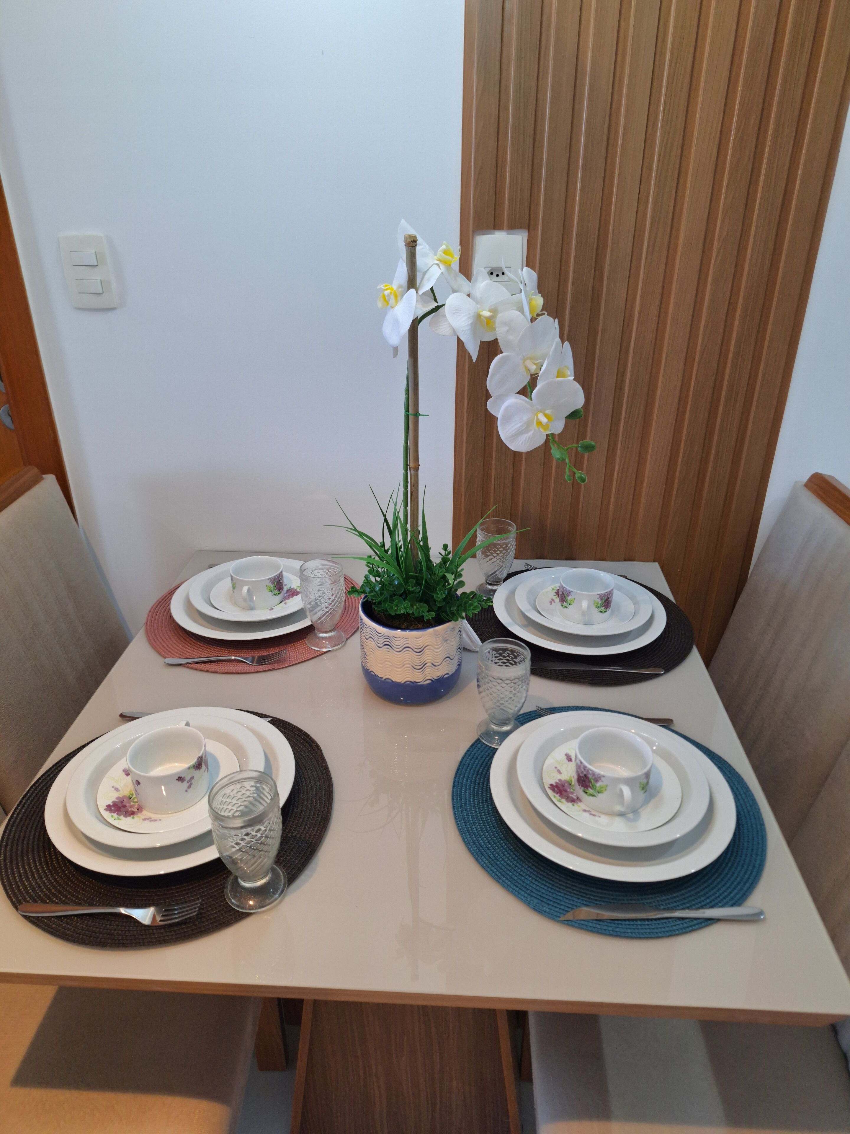 Una mesa de comedor dispuesta con platos, cubiertos y una planta en maceta con flores blancas.