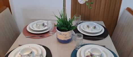 Una mesa de comedor dispuesta con platos, cubiertos y una planta en maceta con flores blancas.