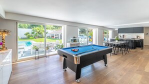 Un salon moderne doté d’une table de billard, d’un coin repas et de grandes fenêtres donnant sur un jardin.