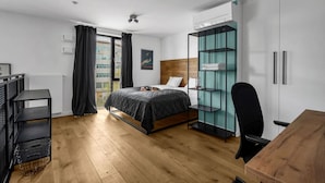 Un dormitorio moderno con piso de madera, una cama con colcha oscura, una silla negra, un escritorio y una estantería adosada a la pared.