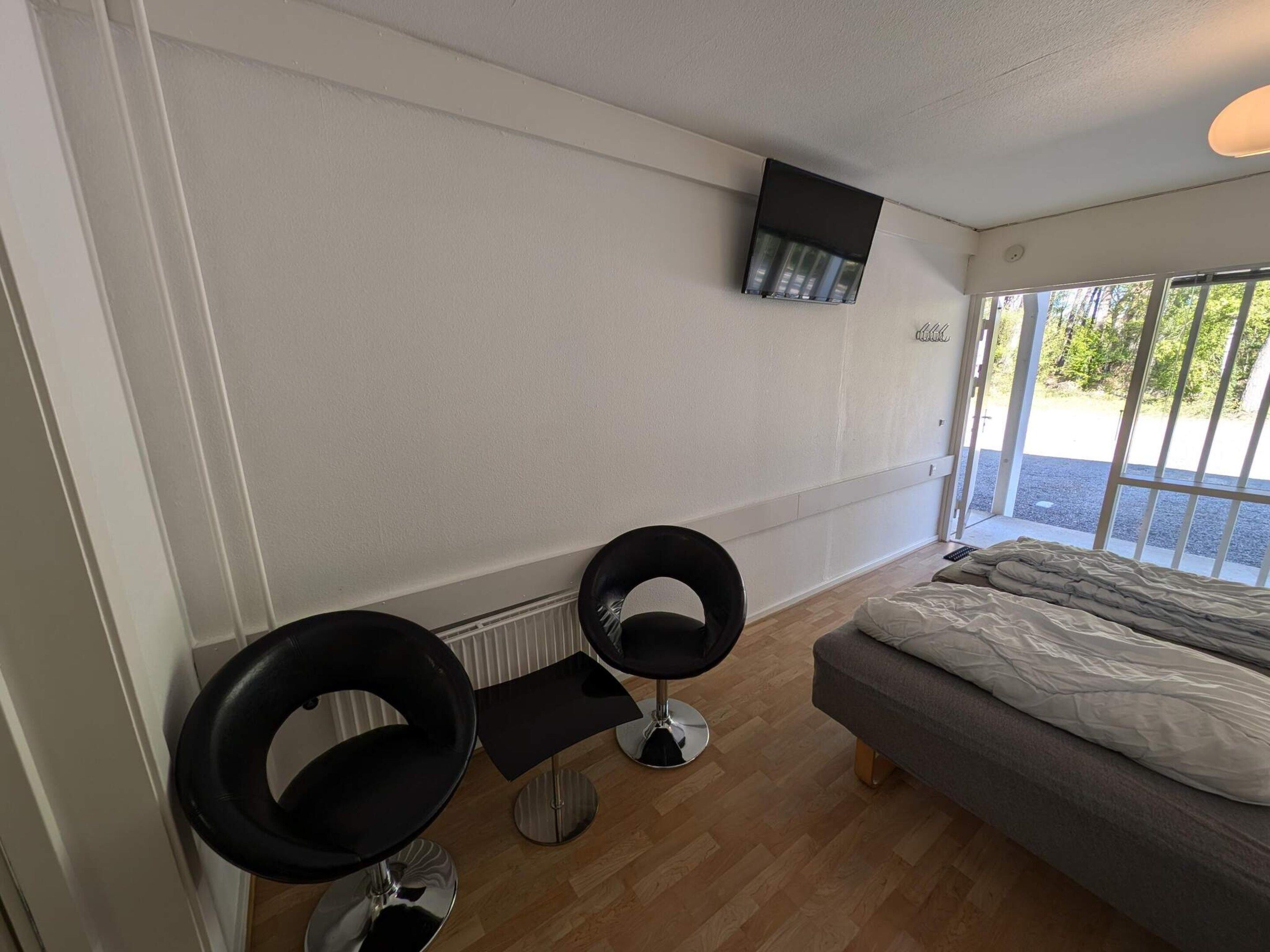 Une pièce comprenant un lit, deux chaises noires, un téléviseur fixé au mur et une fenêtre donnant sur un paysage verdoyant.