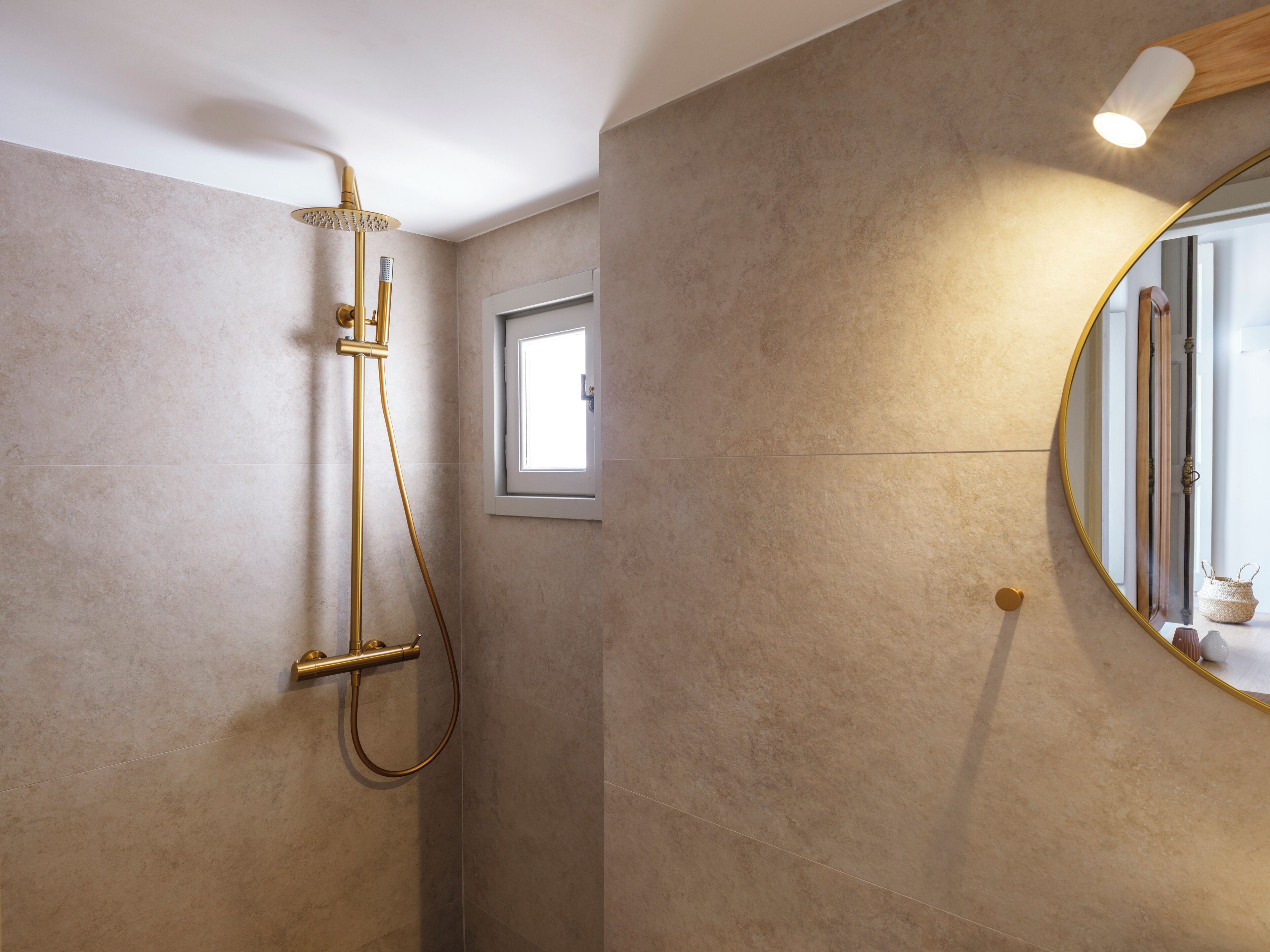 A bathroom with a gold shower system, a small window, and a round mirror with a gold frame.